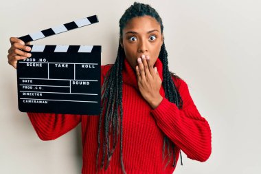 African american woman with braids holding video film clapboard covering mouth with hand, shocked and afraid for mistake. surprised expression 