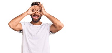 Latin asıllı genç bir adam, günlük beyaz tişört giyiyor dürbün gibi jest yapıyor, dili dışarı çıkarıyor, gözleri parmaklarından bakıyor. çılgın ifade. 