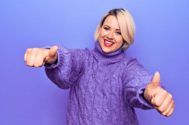 Güzel sarışın ve boyunda bir kadın mor arka plan üzerine balıkçı yaka kazak giyip olumlu el hareketlerini onaylıyor, gülümseyerek ve başarı için mutlu. Kazanan jest.
