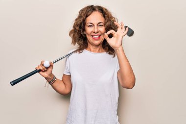 Orta yaşlı güzel sporcu kadın golf oynuyor beyaz arka planda top ve sopayla oynuyor tamam işareti yapıyor, gülümsüyor arkadaşça jestler yapıyor mükemmel bir sembol