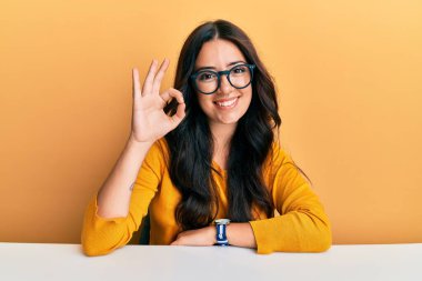 Güzel, esmer, gözlüklü ve rahat elbiseli bir kadın masada oturmuş gülümsüyor ve el ve parmaklarıyla işaret veriyor. başarılı ifade. 