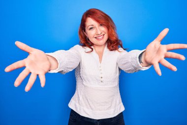 Güzel, kızıl saçlı, zarif gömlekli bir iş kadını mavi arka planda durup kameraya sarılmak için gülümseyerek bakıyor. Mutluluğu kucaklayan neşeli bir ifade..