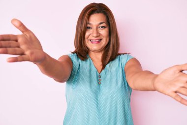 Orta yaşlı esmer, sıradan kıyafetler giyen, kucaklaşmak için kollarını açıp kameraya gülümseyen Latin bir kadın. Mutluluğu kucaklayan neşeli bir ifade.. 