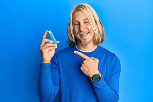 Caucasian young man with long hair holding medical asthma inhaler smiling happy pointing with hand and finger