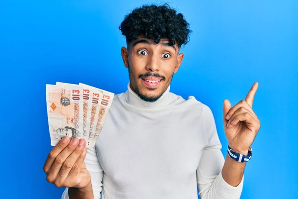 年轻的Arab英俊男子 手里拿着10英镑的英国钞票 高兴地微笑着 手指手画脚地指向旁边 — 图库照片