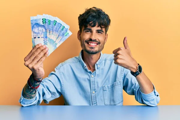 Joven Hispano Sosteniendo Billetes Rand Sudafricanos Sentado Mesa Sonriendo Feliz —  Fotos de Stock