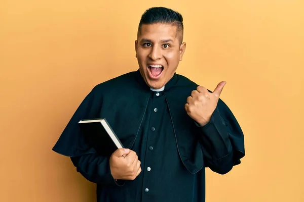 Joven Sacerdote Latino Sosteniendo Biblia Apuntando Con Pulgar Hacia Costado — Foto de Stock