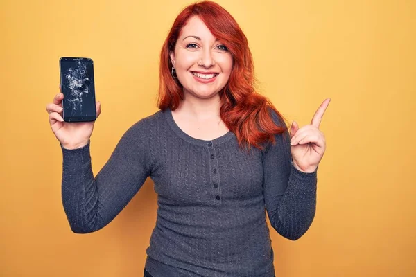 Young beautiful redhead woman holding broken smartphone showing cracked screen smiling happy pointing with hand and finger to the side