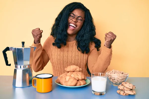 テーブルの上に座っている美しいアフリカの女性は 朝の朝食を食べ 腕を上げて目を閉じて成功に驚いて驚いています — ストック写真