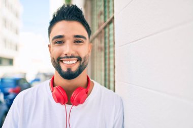 Genç Arap adam, şehrin duvarına dayanmış kulaklık takarak mutlu bir şekilde gülümsüyor..