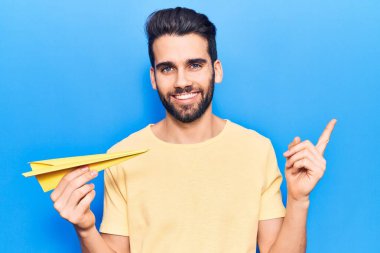 Sarı kağıt uçak tutan sakallı yakışıklı genç adam mutlu bir şekilde elini ve parmağını yana doğru uzattı. 