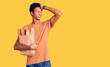 Genç Afro-Amerikan adam elinde kağıt torba, elinde ekmek, gülümsüyor, saçlarına dokunuyor, el hareketi yapıyor, çekici ve modaya uygun poz veriyor. 