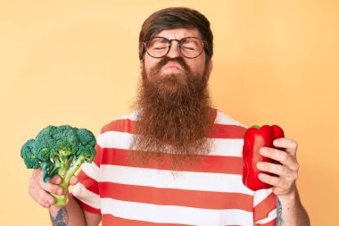 Uzun sakallı, elinde brokoli ve kırmızı biber olan yakışıklı kızıl saçlı bir adam öpüşürken kameraya bakıyor ve sevimli ve seksi. aşk ifadesi. 