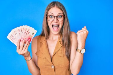 Sarışın kız İsrail şekellerini tutuyor gururla bağırıyor, zaferi kutluyor ve kollarını kaldırarak çok heyecanlanıyor. 