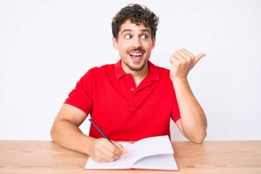 Genç, beyaz, kıvırcık saçlı, defter yazan, masaya oturan, baş parmağını yan tarafa kaldırıp gülümseyen, ağzı açık, mutlu bir adam. 