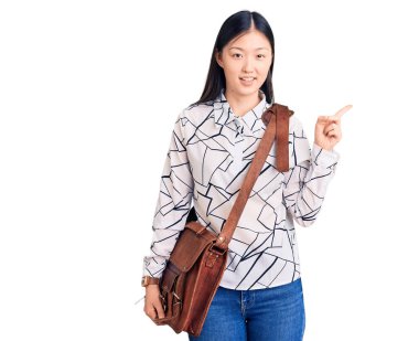 Genç, güzel Çinli bir kadın evrak çantası tutuyor. Gülümsüyor, mutlu bir şekilde elini ve parmağını yana doğru gösteriyor. 