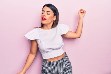 Genç, güzel, esmer, günlük kıyafetler giyen bir kadın izole edilmiş pembe arka planda dikiliyor.