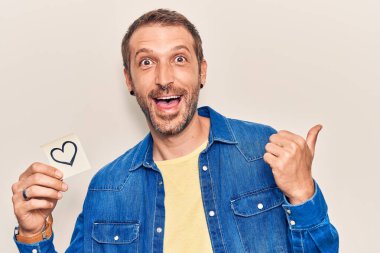 Genç, yakışıklı bir adam, kalbi tutuyor. Parmağını yana doğru kaldırıyor. Ağzını açıp gülümsüyor. 