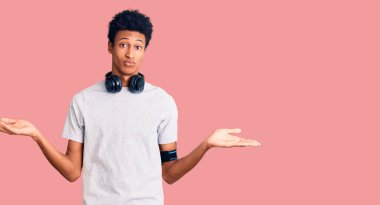 Spor kıyafetleri giyen, kulaklık takan, elleri ve kolları havada, kafası karışmış genç Afro-Amerikan bir adam. şüphe kavramı. 