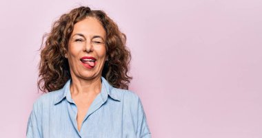 Orta yaşlı, güzel bir kadın pembe arka planda dikilmiş kameraya seksi bir ifadeyle bakıyor, neşeli ve mutlu bir yüzle..