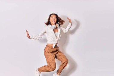 Genç, güzel Çinli kadın mutlu gülümsüyor. Yüzünde gülümsemeyle zıplıyor, beyaz arka planda kulaklık kullanarak müzik dinliyor.