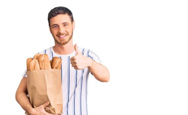 Elinde ekmek torbası olan yakışıklı genç adam mutlu ve pozitif gülümsüyor, başparmağıyla mükemmel ve onay işareti yapıyor. 