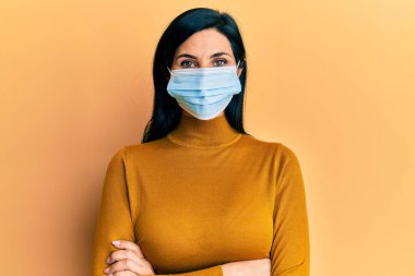 Tıbbi maske takan, kollarını kavuşturup kameraya gülümseyen beyaz bir kadın. pozitif kişi. 