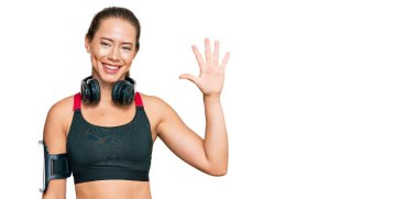 Güzel sarışın kadın spor kıyafetleri giyiyor ve kulaklık takıyor. Beş numarayı gösterip işaret ederken kendine güvenen ve mutlu bir şekilde gülümsüyor.. 