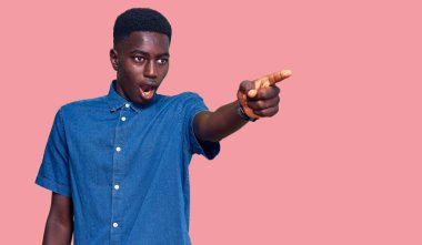 Genç, Afro-Amerikalı, günlük giysiler giymiş, parmaklarını göstererek şaşırmış, ağzı açık, şaşırmış, ön tarafta bir şey var. 