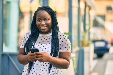 Genç Afrikalı Amerikalı kadın şehirde akıllı telefon kullanarak mutlu bir şekilde gülümsüyor..