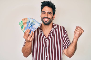 Sakallı yakışıklı İspanyol adam Güney Afrika rand banknotları tutuyor gururla bağırıyor, zaferi kutluyor ve başarıyı kollarını kaldırarak kutluyor. 
