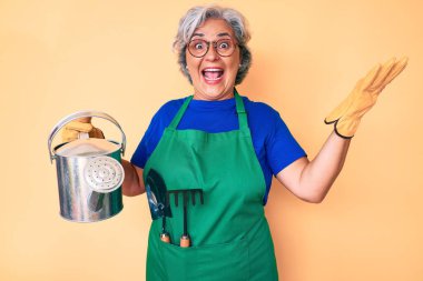 Bahçıvan önlüğü ve eldiven takan yaşlı, beyaz saçlı kadın zaferi mutlu bir gülümsemeyle ve ellerini kaldırarak kutluyor. 