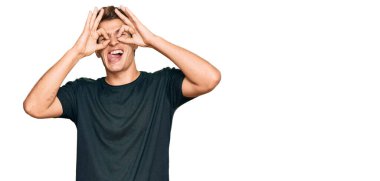 Yakışıklı, günlük kıyafetler giyen beyaz adam dürbün gibi jest yapıyor, dili dışarı çıkarıyor, gözleri parmaklarından bakıyor. çılgın ifade. 