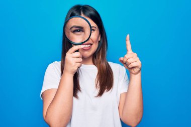 Genç, güzel esmer kadın, izole edilmiş mavi arka plan üzerinde büyüteç kullanıyor. Bir fikir ya da soru işareti ile gülümsüyor.