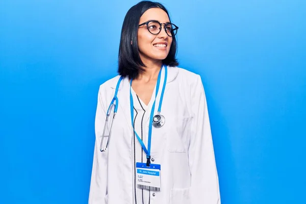 Young beautiful latin woman wearing doctor stethoscope and id card looking away to side with smile on face, natural expression. laughing confident.
