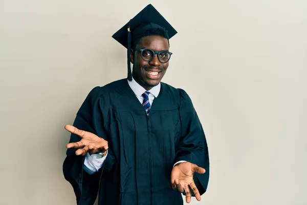 Handsome black man wearing graduation cap and ceremony robe smiling cheerful with open arms as friendly welcome, positive and confident greetings