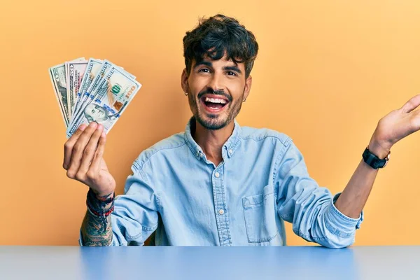 Joven Hombre Hispano Sosteniendo Dólares Sentado Mesa Celebrando Logro Con —  Fotos de Stock