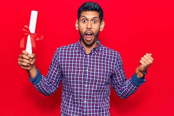 Mezuniyet diploması olan genç latin bir adam baş parmağını yana kaldırmış, ağzı açık gülümsüyor. 