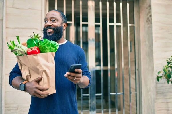 Sakallı Afrikalı Amerikalı adam elinde süpermarketten alınmış kağıt torbalarla akıllı telefon kullanıyor.