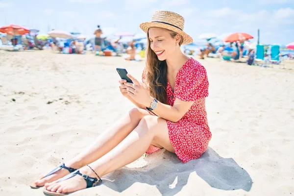 Genç sarışın kadın tatilde. Kumsalda kumsalda oturmuş akıllı telefon kullanıyor.