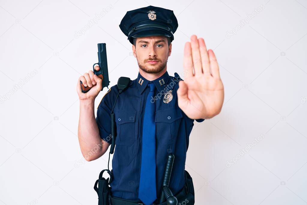 Young caucasian man wearing police uniform holding gun with open hand doing stop sign with serious and confident expression, defense gesture 
