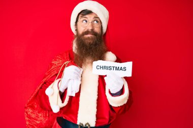 Uzun sakallı yakışıklı kızıl saçlı, Noel Baba kostümü giyen, elinde Noel mesajı tutan, gülümseyen ve kafasını çeviren bir adam.. 