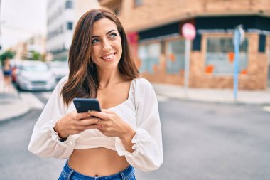 Genç İspanyol kadın şehirde akıllı telefon kullanarak mutlu bir şekilde gülümsüyor..
