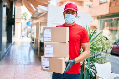 Üniforma giyen ve Coronavirus koruması takan genç bir teslimatçı. Sağlık maskesi. Kasaba sokaklarında karton paketler tutuyor..