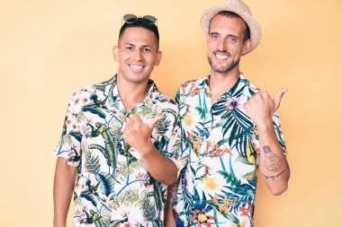 Yazlık şapkalı ve Hawaii tişörtlü iki genç eşcinsel el ve baş parmakları havada, kendinden emin bir şekilde arkasını gösteriyorlar. 