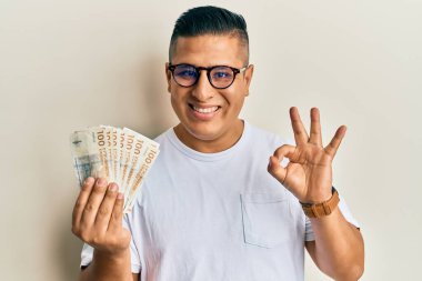 Genç latin adam elinde 100 Danimarka kronu tutuyor. Parmaklarıyla imza atıyor, gülümsüyor ve mükemmel bir sembol oluşturuyor. 