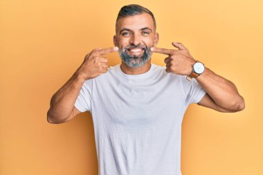 Orta yaşlı, beyaz tişört giyen, gülümseyen, parmaklarıyla ve ağzıyla işaret eden yakışıklı bir adam. diş sağlığı kavramı. 