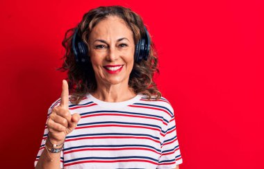 Orta yaşlı esmer kadın kırmızı arka planda kulaklık takarak müzik dinliyor. Bir fikir veya soruyla gülümsüyor ve mutlu bir yüzle işaret ediyor.