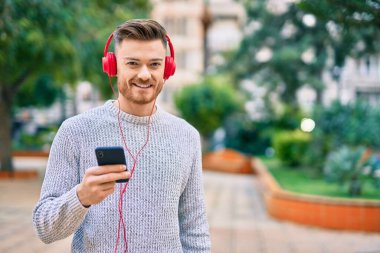 Parkta akıllı telefon ve kulaklık kullanarak gülümseyen beyaz bir genç..