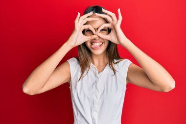 Genç esmer kadın, günlük kıyafetler ve gözlük takıyor dürbün gibi jest yapıyor, dili dışarı çıkarıyor, gözleri parmaklarından bakıyor. çılgın ifade. 